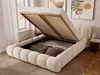 A cream-colored upholstered bed with its storage compartment open, revealing pillows and bedding inside. The bed is in a room with artwork on the wall and a window with curtains. Produktbild-Vorschau