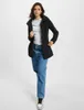 A woman is standing and posing in a studio setting, wearing a black jacket and blue jeans. Produktbild-Vorschau