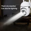 A white security camera is mounted indoors, emitting a bright beam of light. The background shows a dimly lit living room with furniture. Podgląd zdjęcia produktu