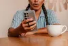 A person holding a smartphone with a ring stand attached, sitting at a table with a cup of coffee. Produktbild-Vorschau
