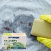 A bar of Heitmann Gallseife soap is being held by a yellow gloved hand over a stained piece of fabric, with the product packaging visible in the foreground. Produktbild-Vorschau