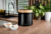 A black appliance with a wooden lid sits on a wooden countertop, next to two white bowls of spices. In the background, a kitchen sink and plants are visible. Voir les images du produit