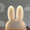 A bunny ear-shaped night light is mounted on a gray wall above a wooden surface. Other wooden cutouts of a house, a car, and a hedgehog are also visible on the wall. Produktbild-Vorschau
