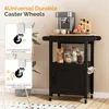 A black kitchen cart with a wooden top, featuring a coffee maker, cups, and fruit on top, and jars of food on a shelf. The cart has a cabinet door and hooks for mugs. Insets show close-ups of the caster wheels. Podgląd zdjęcia produktu