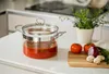 A glass cooking pot filled with tomato sauce sits on a white countertop next to some fresh ingredients. Náhled obrázku produktu
