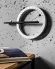 A modern, circular calendar is mounted on a textured concrete wall. A book lies on a wooden table in the foreground. Produktbild-Vorschau