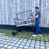 A man is pushing a black and white folding scaffold with two levels on a paved area with grass. Anteprima dell’immagine prodotto