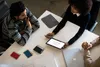Three people are gathered around a white table, looking at a tablet and a laptop. Produktbild-Vorschau