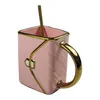 A pink ceramic mug with a gold handle and trim, designed to resemble a quilted handbag. A gold spoon is placed inside the mug. Produktbild-Vorschau