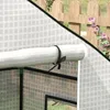 A close-up view of a rolled-up cover for a greenhouse, secured with a dark green tie. The cover has a white background with a subtle grid pattern. In the background, a wooden fence and pink flowers are visible. Voir les images du produit