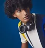 A young man with curly hair wearing headphones around his neck and a striped shirt against a blue background. Produktbild-Vorschau