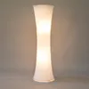 A tall, white, hourglass-shaped floor lamp is shown against a plain, light gray background. Produktbild-Vorschau