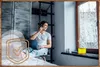 A man is sitting on a chair next to a window, talking on the phone and working on his laptop. There is a humidifier on the windowsill. Produktbild-Vorschau