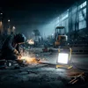 A welder is working in a dimly lit industrial setting, with a portable work light illuminating the foreground. Anteprima dell’immagine prodotto