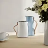 Two thermoses and a coffee cup are arranged on a wooden table with a vase of flowers in the background. Produktbild-Vorschau