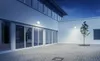 An outdoor courtyard at dusk with a modern building and a single tree. Produktbild-Vorschau