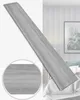 A single plank of wood-look material is shown diagonally against a blurred background of a room with a bed and artwork. Anteprima dell’immagine prodotto