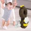 A baby is learning to walk with the help of an adult, and a bee-shaped safety backpack is placed next to them. Podgląd zdjęcia produktu