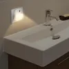 A white LED night light with a motion sensor is plugged into a wall outlet next to a bathroom sink. Produktbild-Vorschau