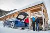 A couple loading luggage into the trunk of a blue electric car in front of a wooden cabin in the snow. Produktbild-Vorschau