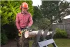 A man wearing safety gear is using a chainsaw to cut a log in a backyard. Anteprima dell’immagine prodotto