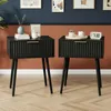 Two black nightstands with fluted drawers and gold handles are shown in a room with dark blue and orange walls and a wooden floor. Produktbild-Vorschau