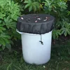 A white rain barrel with a black mesh cover is placed outdoors amongst green foliage and grass. Produktbild-Vorschau