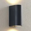 A black, semi-circular wall lamp mounted on a textured gray wall, emitting light upwards and downwards. Náhled obrázku produktu