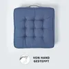 A blue tufted seat cushion is shown from a top-down perspective against a light gray background. A circular graphic with a hand sewing and the text "VON HAND GESTEPPT" is in the bottom left corner. Náhled obrázku produktu