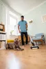 A man is vacuuming a living room floor while his dog lies nearby. Anteprima dell’immagine prodotto