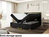 A bedroom scene with a black boxspring bed with its storage compartment open, revealing the mattress and topper. The bed has gold accents and is placed on a rug. A vase with dried flowers is visible on the left, and a nightstand with a lamp is on the right Produktbild-Vorschau
