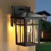 An outdoor wall lamp is mounted on a wall, with a house and garden visible in the background. Produktbild-Vorschau