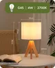 A bedside table lamp with a wooden base and a white fabric shade, placed on a white table next to a chair and a book. Produktbild-Vorschau