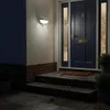 A dark blue front door with glass panels and a gold knocker and mail slot, illuminated by a white wall-mounted outdoor light. Steps leading up to the door are visible, with fallen leaves scattered on them and the surrounding dark stone patio. Potted plants Produktbild-Vorschau