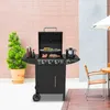 A black gas grill with food cooking on it, placed outdoors next to a potted plant and a table with chairs. Produktbild-Vorschau