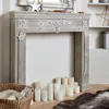 A fireplace mantel with decorative carvings and a distressed wood finish, surrounded by candles and a basket with a fluffy item inside. Anteprima dell’immagine prodotto
