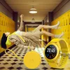A woman is floating in a school hallway with yellow lockers. A yellow smartwatch and a round yellow case are in the foreground. Voir les images du produit