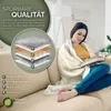 A woman is sitting on a couch wrapped in a beige blanket, reading a book. A diagram showing the layers of the blanket is on the left. Voir les images du produit