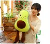 A woman is holding a plush avocado toy in a room with wooden furniture and plants. Produktbild-Vorschau