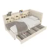 A beige upholstered bed with a built-in shelf unit at the headboard and two open drawers underneath, displaying various items. The bed is shown from a slightly elevated, diagonal angle. Náhled obrázku produktu