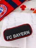 A black pencil case with 'FC BAYERN' written on it, placed on an open notebook with math problems. A red and blue striped scarf with the FC Bayern logo is visible in the background. Anteprima dell’immagine prodotto