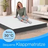 A woman is sitting on a foldable mattress in a living room setting. Produktbild-Vorschau