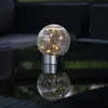 A solar-powered LED light globe with a cracked glass effect sits on a reflective surface, with blurred outdoor furniture in the background. Produktbild-Vorschau