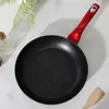 A frying pan with a red handle is placed on a white surface, with some greenery and a glass bottle in the background. Voir les images du produit