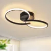 A modern black ceiling light with two intertwined rings, illuminated with warm white light, mounted on a white ceiling. Voir les images du produit