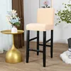 A beige bar stool cover is shown on a bar stool, with a small gold side table and a vase of flowers to the left, and a plant to the right. Produktbild-Vorschau
