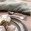 A table setting with cutlery, a napkin, and a plate, arranged on a wooden surface with a light green cloth draped in the background. Produktbild-Vorschau