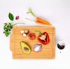 A bamboo cutting board with various fresh vegetables and herbs arranged on it, set against a white background. Produktbild-Vorschau