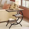 A narrow, C-shaped side table with three shelves is placed next to a sofa, with a vintage telephone and a lamp on the top shelf. Voir les images du produit