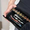 A person's hand is holding open a black wallet, revealing euro banknotes and coins organized in compartments. A smartphone is also visible inside the wallet. Podgląd zdjęcia produktu